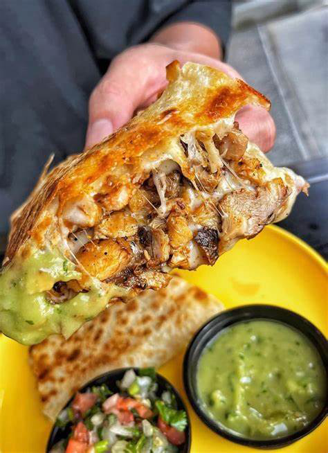 A hand holds a grilled burrito showing layers of melted cheese and meat, next to a yellow plate with salsa and dip.