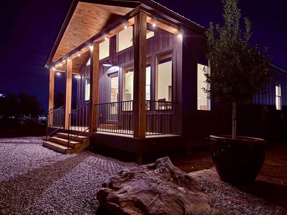 Modern dark-sided cabin at night with a covered porch, wood pillars, glowing string lights, and a gravel yard.