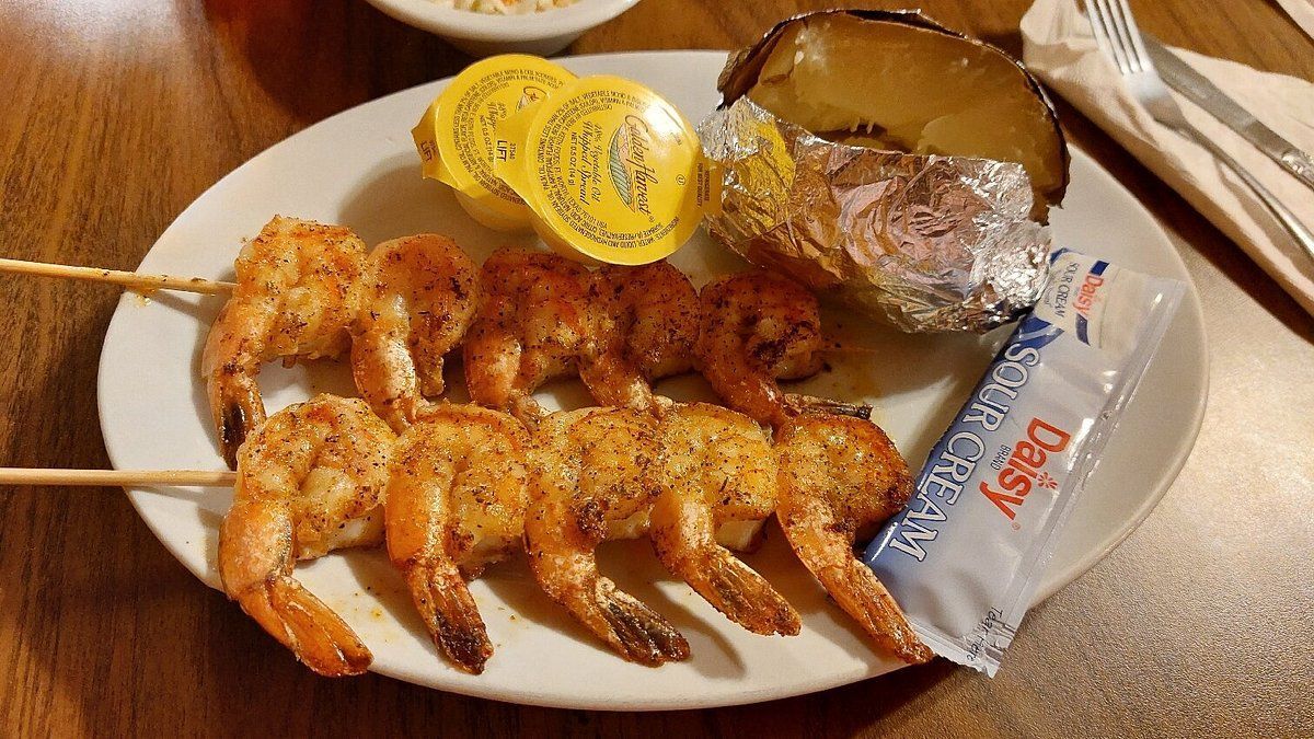 A meal featuring two skewers of grilled, seasoned shrimp, a baked potato in foil, butter cups, and a packet of sour cream.