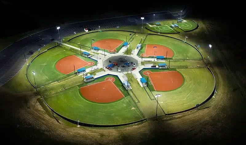 An aerial night view of a four-field baseball complex with illuminated diamonds radiating from a central hub.