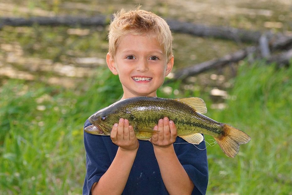 A smiling child holds a caught green and black bass fish in front of a grassy outdoor background.