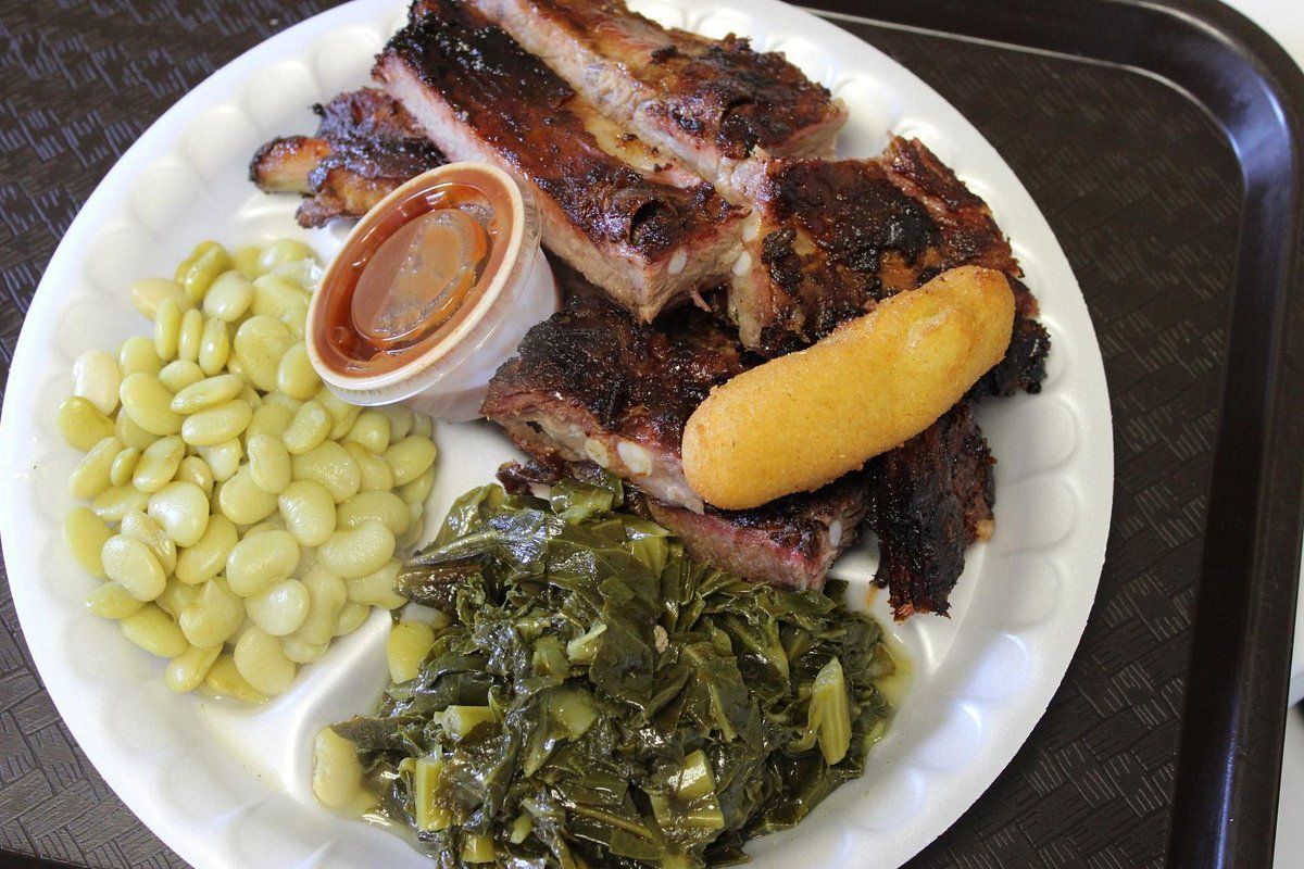 A white plate holding barbecue pork ribs, a side of lima beans, cooked greens, a small cup of sauce, and a hush puppy.