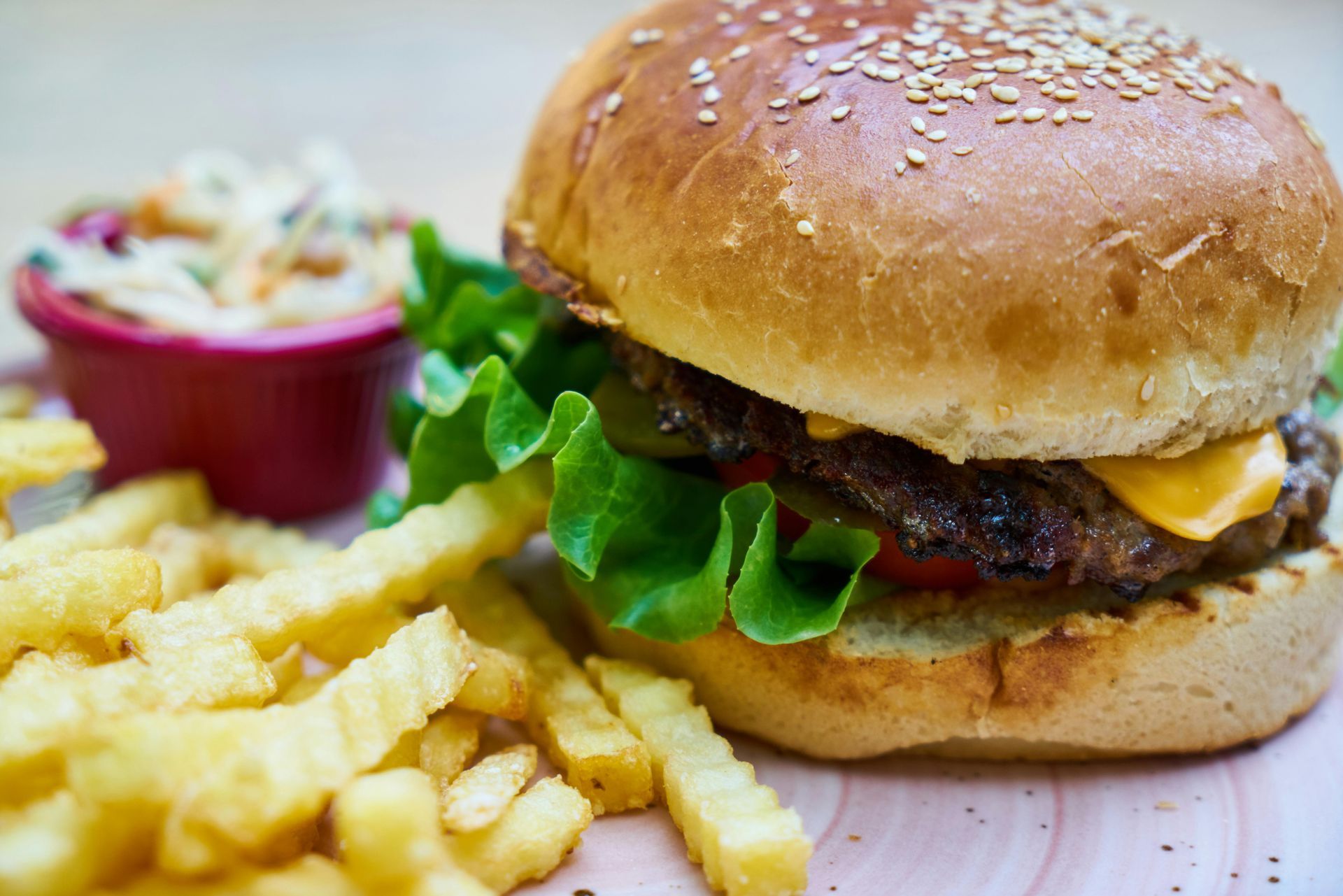 A sesame seed bun burger with lettuce and cheese, served with crinkle-cut fries and a side of coleslaw on a pink plate.