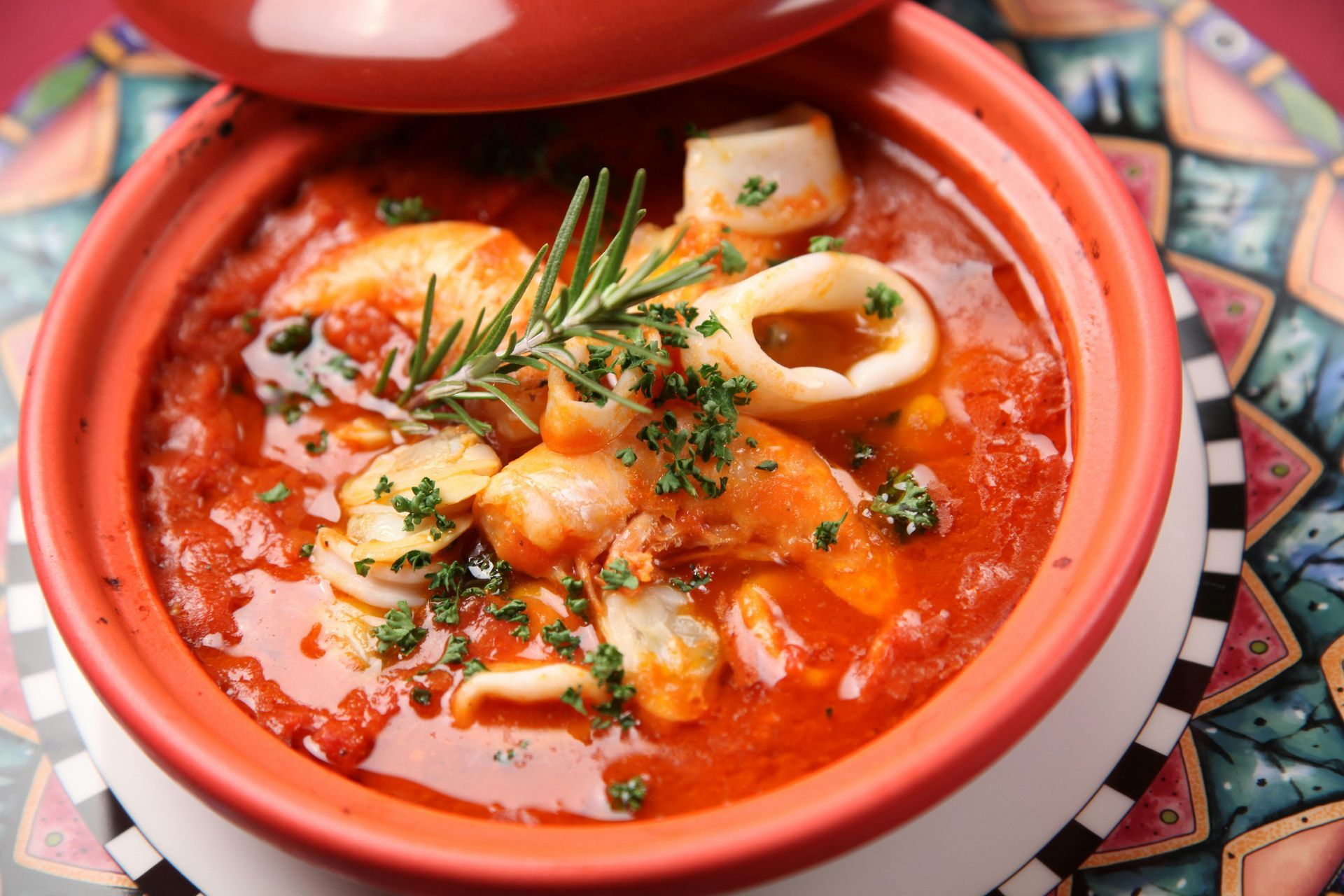 A red clay pot filled with seafood stew, including shrimp and calamari in a tomato sauce, topped with fresh rosemary.