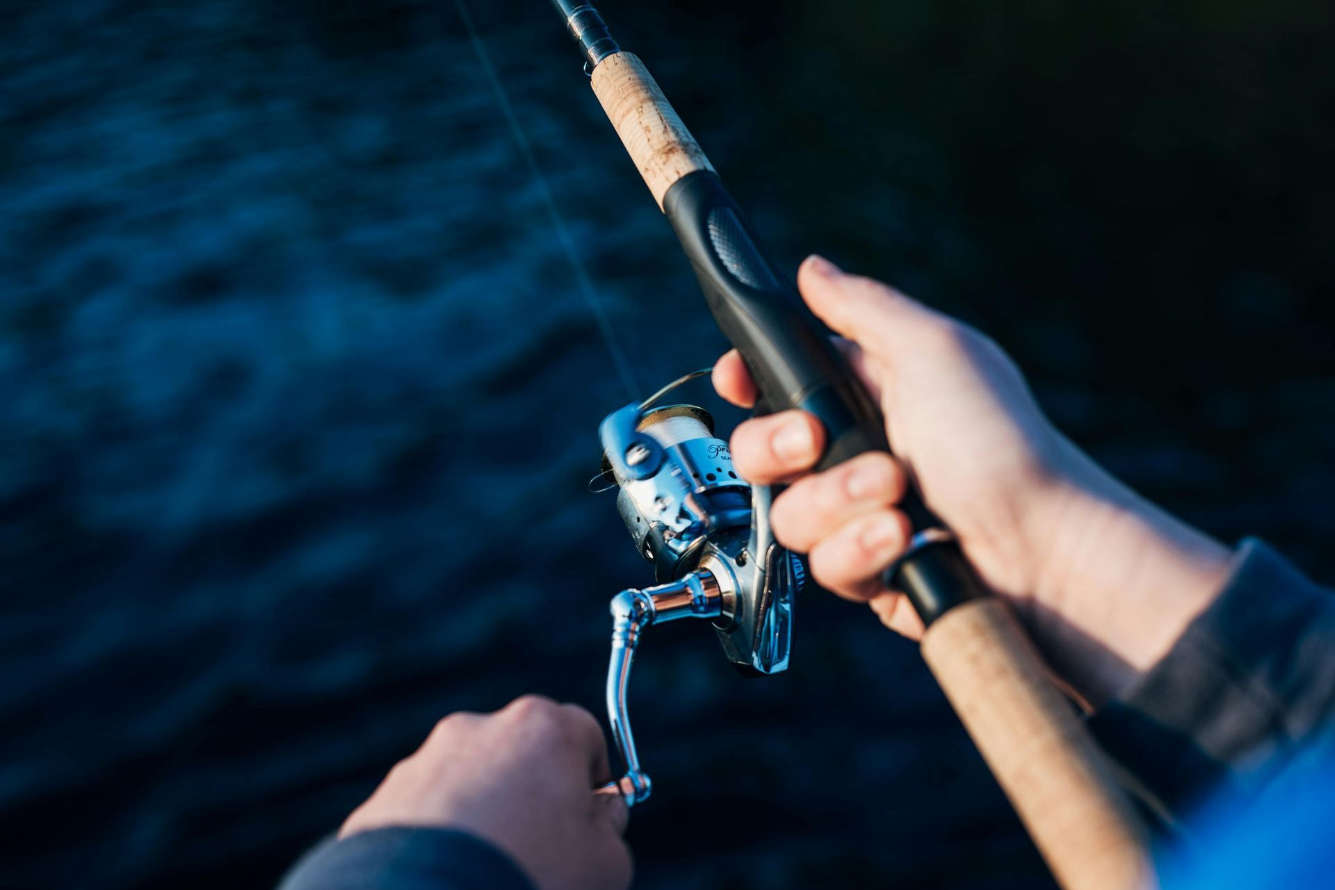 A person holds a spinning fishing rod and reel over dark water.
