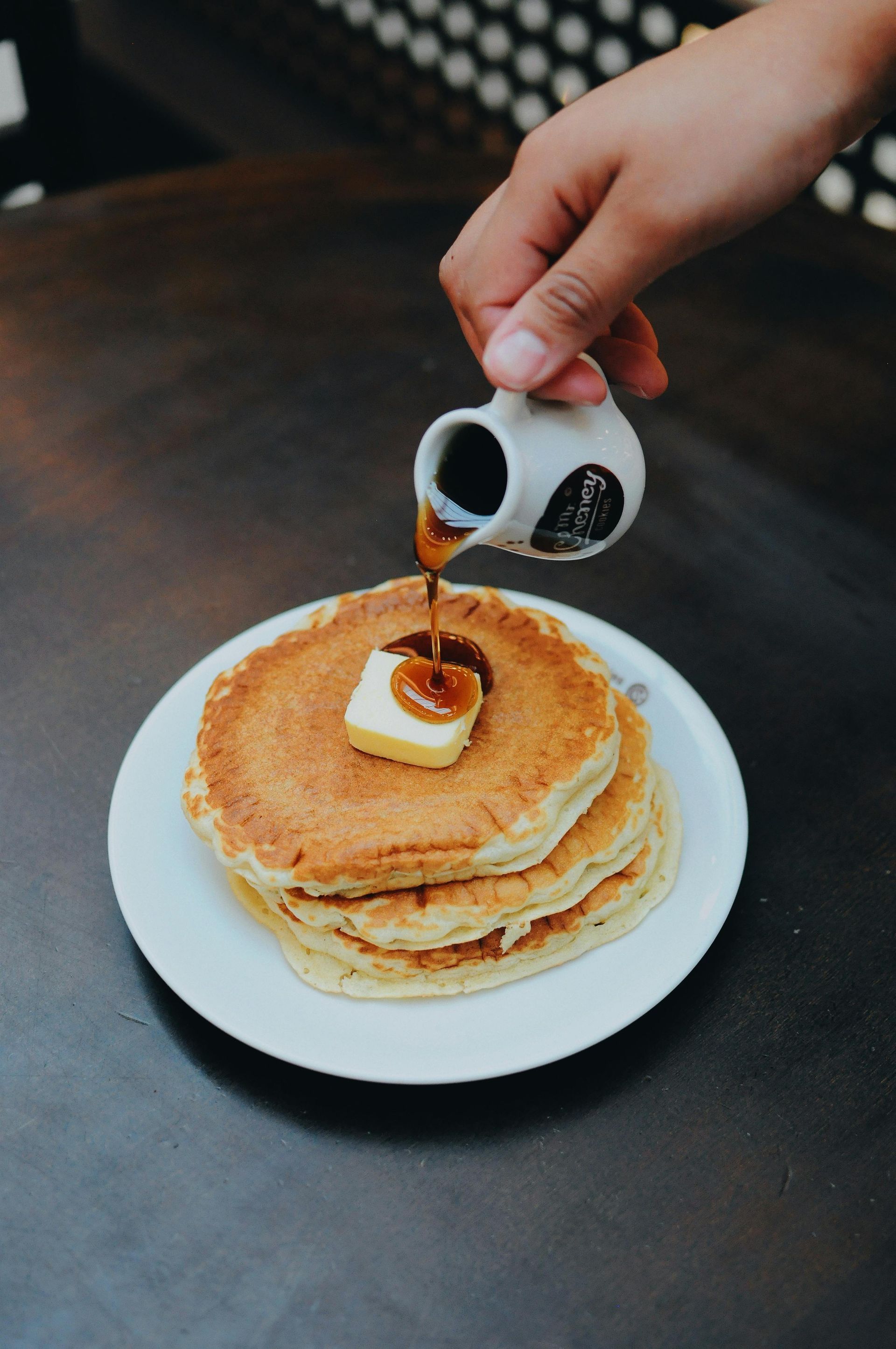 A hand pours maple syrup from a small white pitcher onto a stack of pancakes topped with a pat of butter on a plate.