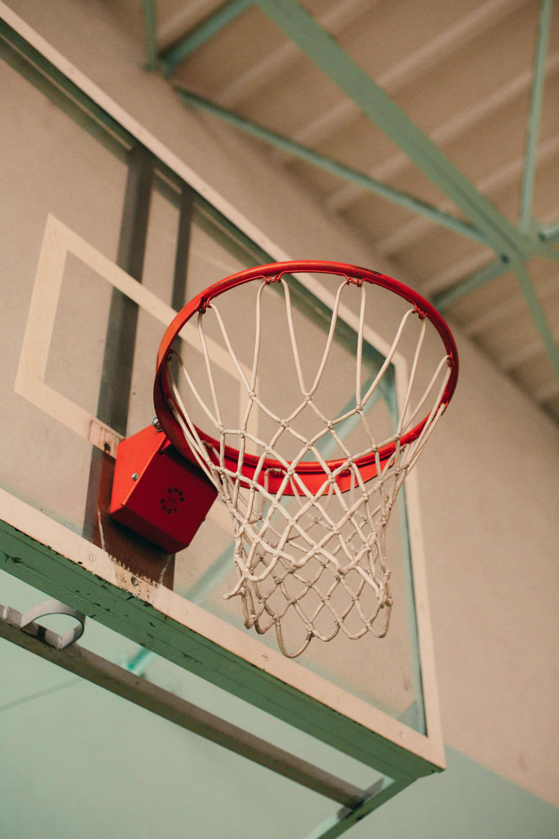A red basketball hoop with a white net mounted on a backboard in an indoor gymnasium.