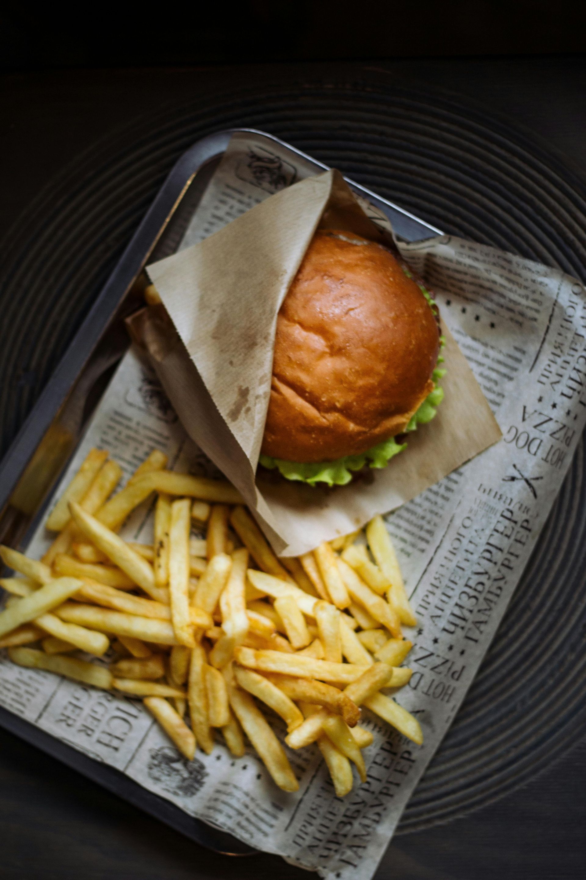 A burger wrapped in paper and a pile of golden-brown french fries on a tray lined with newsprint paper.