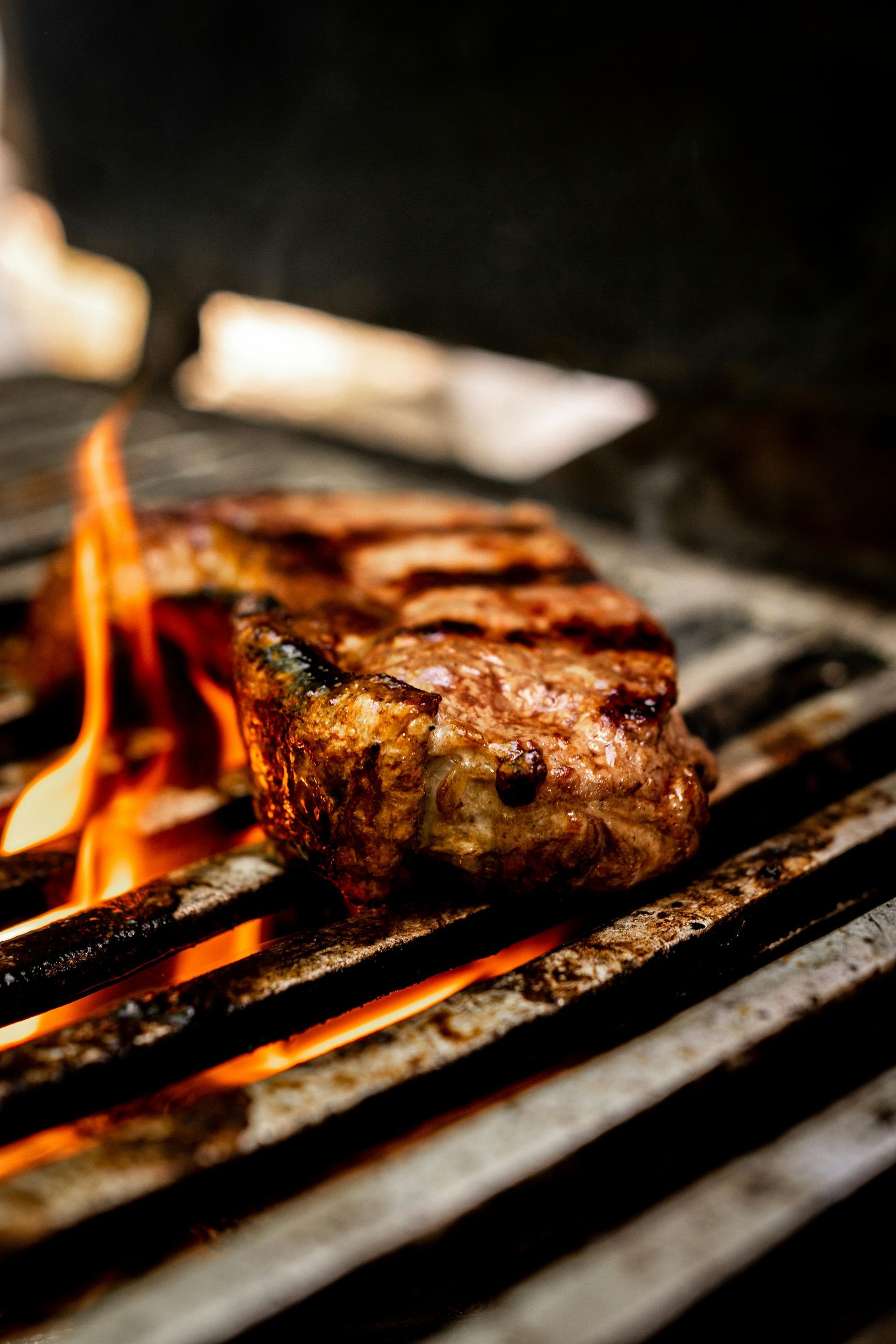 A thick, browned steak searing on hot metal grill grates with visible orange flames licking up the side.