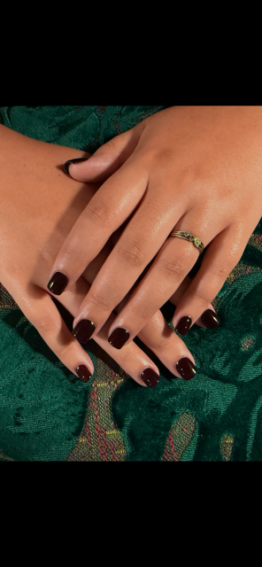 Hands with dark red nail polish, resting on green fabric, with a gold ring.