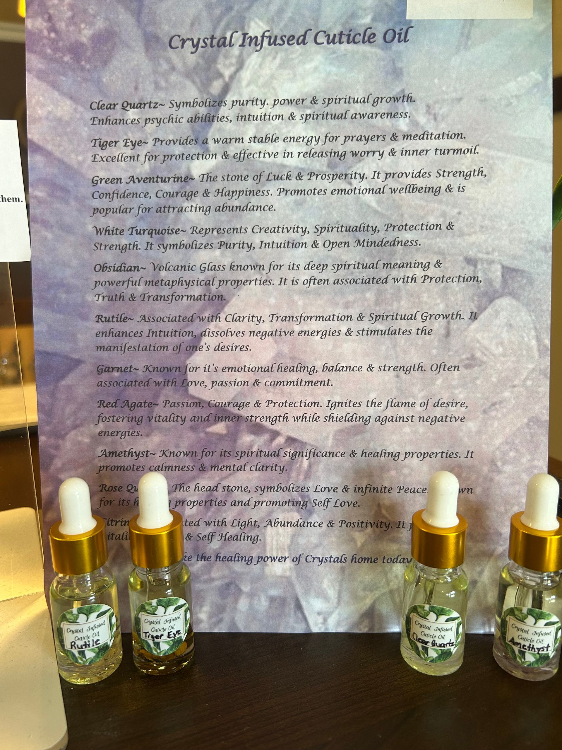 Four small bottles of cuticle oil with a sign describing crystal-infused oil. The sign is purple and text heavy.