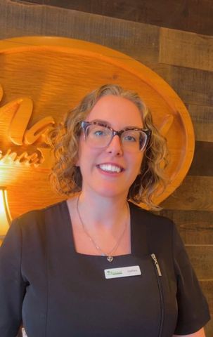 Woman with curly hair wearing glasses, smiling, and wearing a name tag, standing in front of a wooden sign.