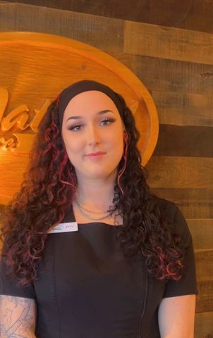 A young woman with long curly hair, a black headband, and a nametag, smiles in front of a wooden backdrop.
