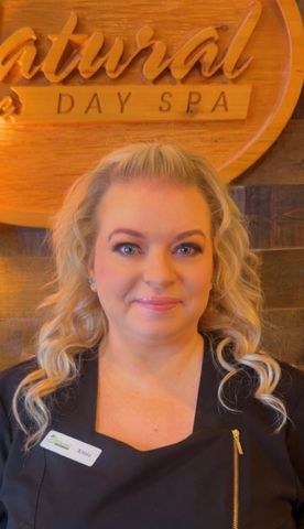 Woman with blonde hair, wearing a black top, smiling at Natural Day Spa. The background is wood.