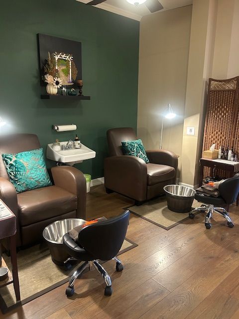 Cozy nail salon with two brown leather chairs, foot basins, and a dark green accent wall.