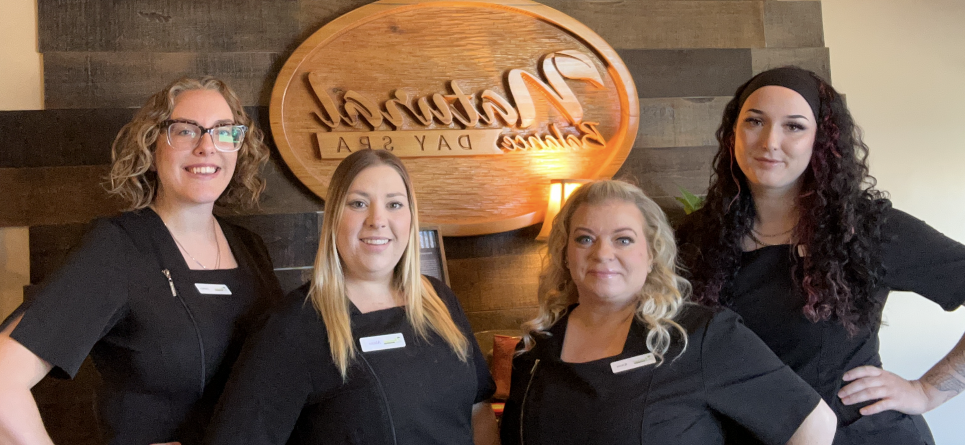 Four women in black shirts smile in front of a wooden sign that says 
