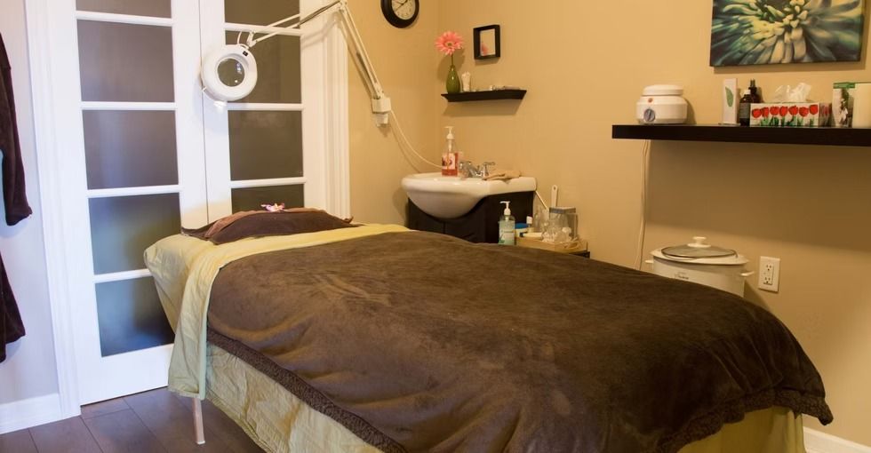 A spa treatment room with a massage table covered in a brown blanket, near a sink and shelf with supplies.