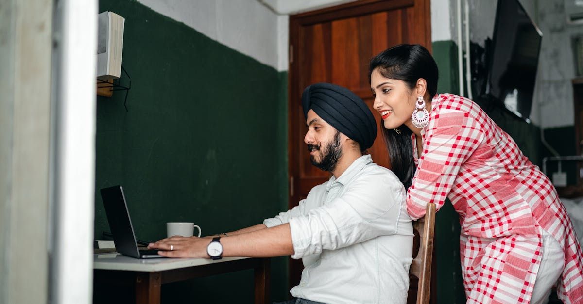 A woman is standing next to a man using a laptop computer.
