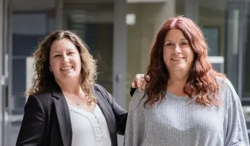 Two smiling people standing side-by-side, one in a black blazer and one in a grey textured top, against a blurred background.