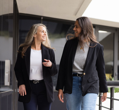 Two people in dark blazers walk outdoors, smiling and conversing while looking toward each other.