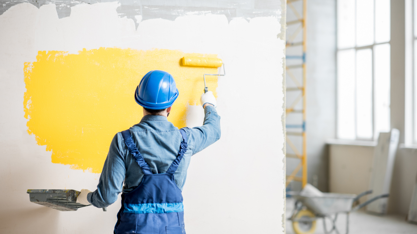 Painter in blue overalls and hard hat, using a paint roller to apply yellow paint to a white wall.