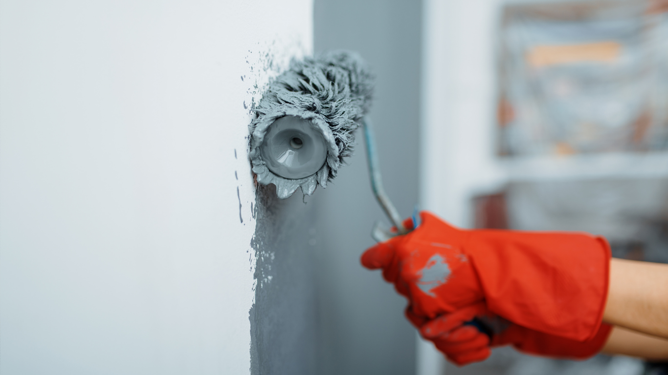 A person in red gloves paints a white wall with a gray paint roller.