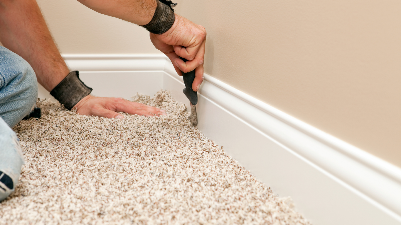 Person using a utility knife to trim carpet along a baseboard.