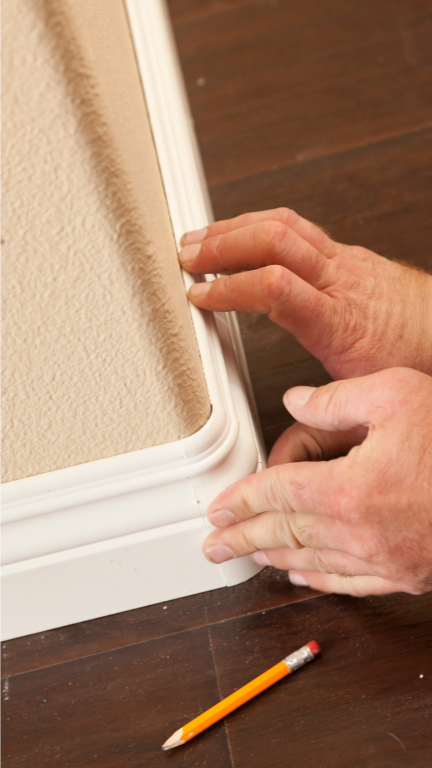 Hands measuring baseboard against carpet and wood flooring with a pencil.