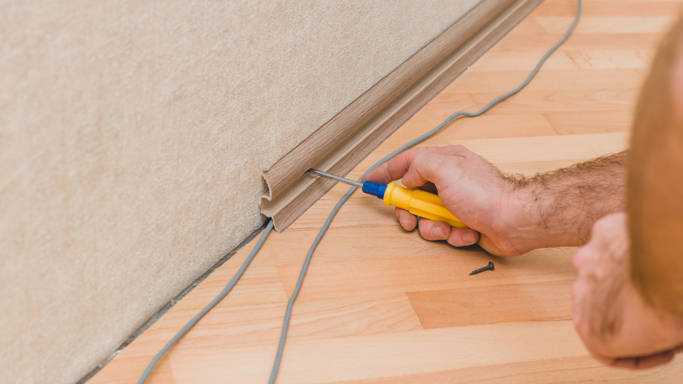 Person using a yellow tool to place grey wires near a baseboard on a wooden floor.