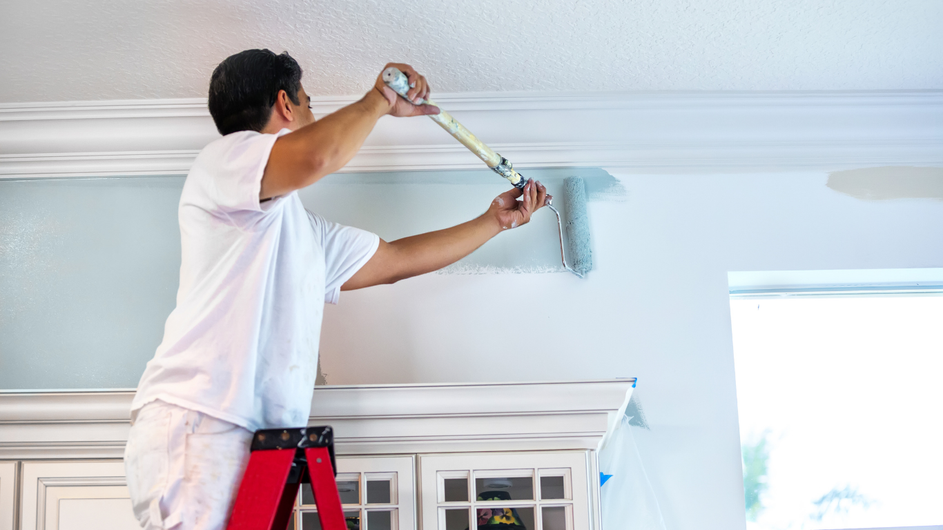 Person paints wall blue with a roller, standing on a red ladder in a room with white cabinets.