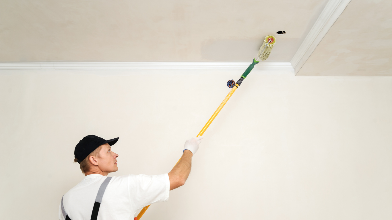 Painter on a ladder using a roller to paint a white ceiling near crown molding.