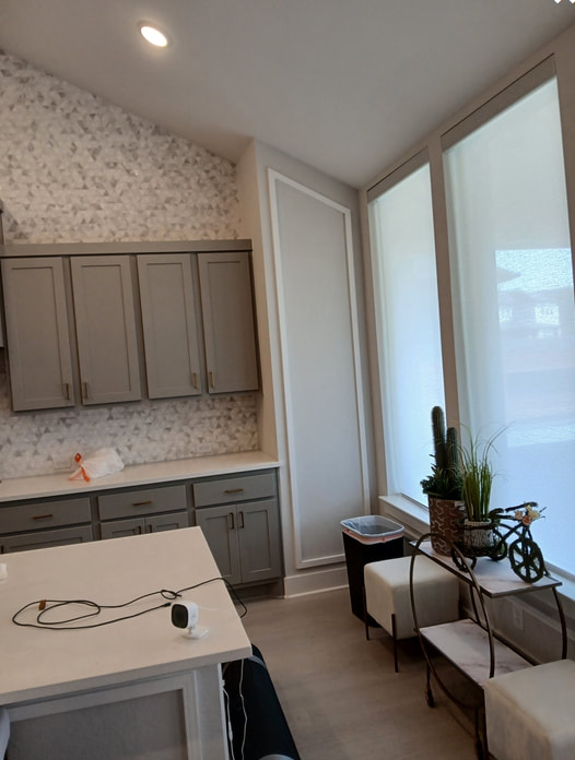 Kitchen with gray cabinets, white countertop, large windows with shades, and a small table with plants.