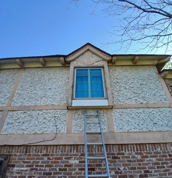 Ladder propped against stucco wall with window, tan beams, and brick base under blue sky.