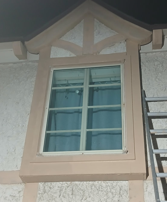 Window with light-colored trim and decorative upper frame on a stucco building, a ladder to the right.
