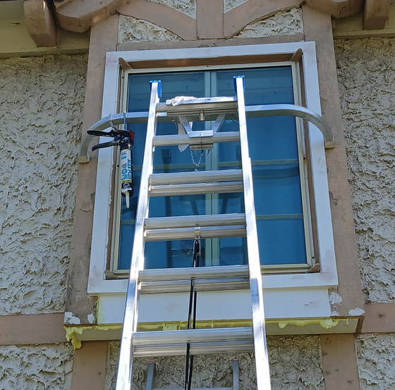 Ladder propped against a window with sealant gun clamped to a curved metal support. Exterior stucco wall.