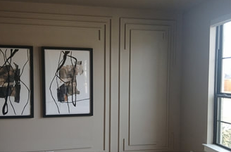 Two abstract framed art pieces hang on a tan wall with decorative molding, next to a window.