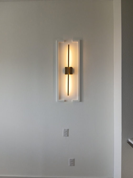Modern wall sconce with a vertical light fixture on a white wall, near a light switch and electrical outlet.