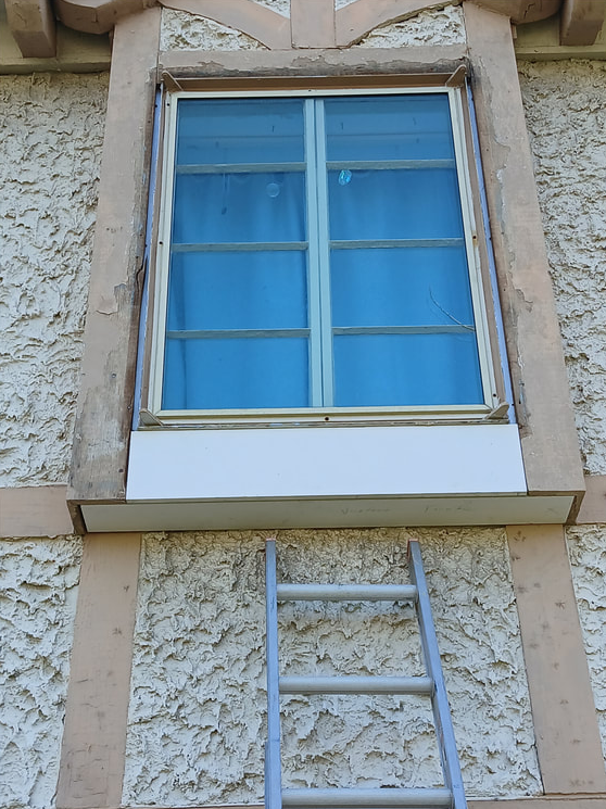 A weathered window with blue-tinted glass, set in a stucco wall, and a ladder beneath it.