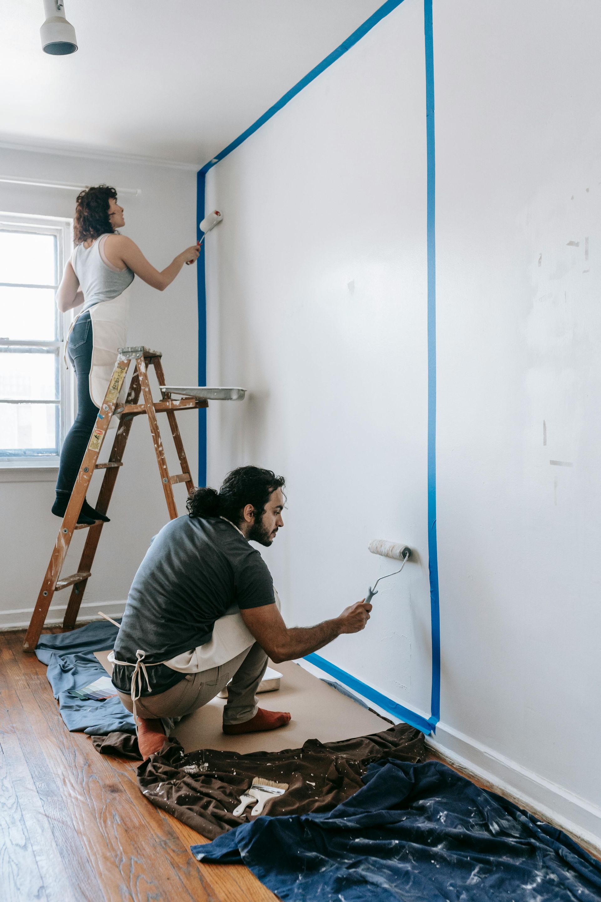 Couple painting a wall with blue tape borders, one on a ladder, indoors.