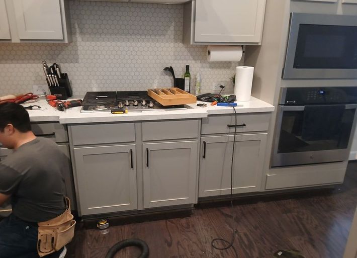 Man working on kitchen cabinets, near stove and oven. Grey cabinets, white countertops and backsplash.