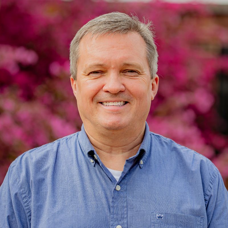 A man in a blue shirt is smiling in front of pink flowers.