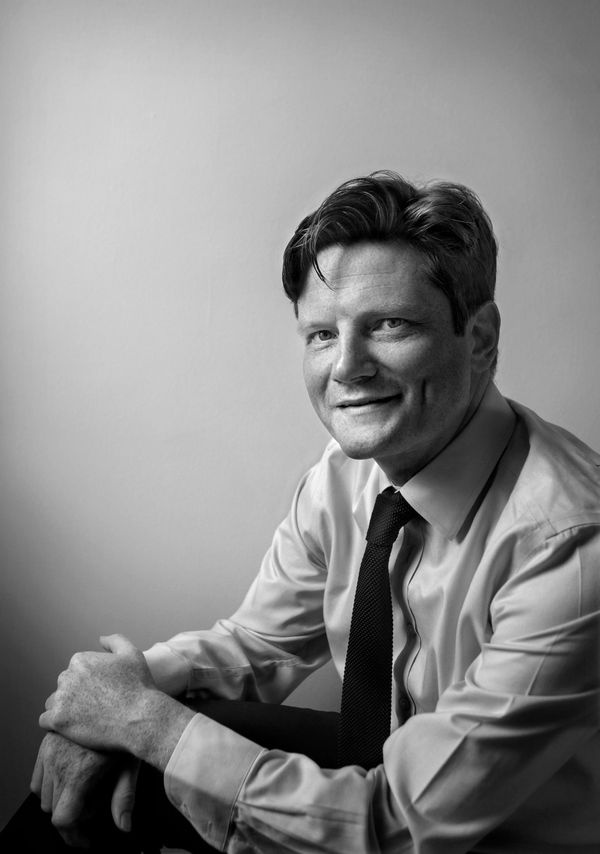 A man with fair skin and thinning hair, wearing a tie and dress shirt, smiles while looking toward the viewer.