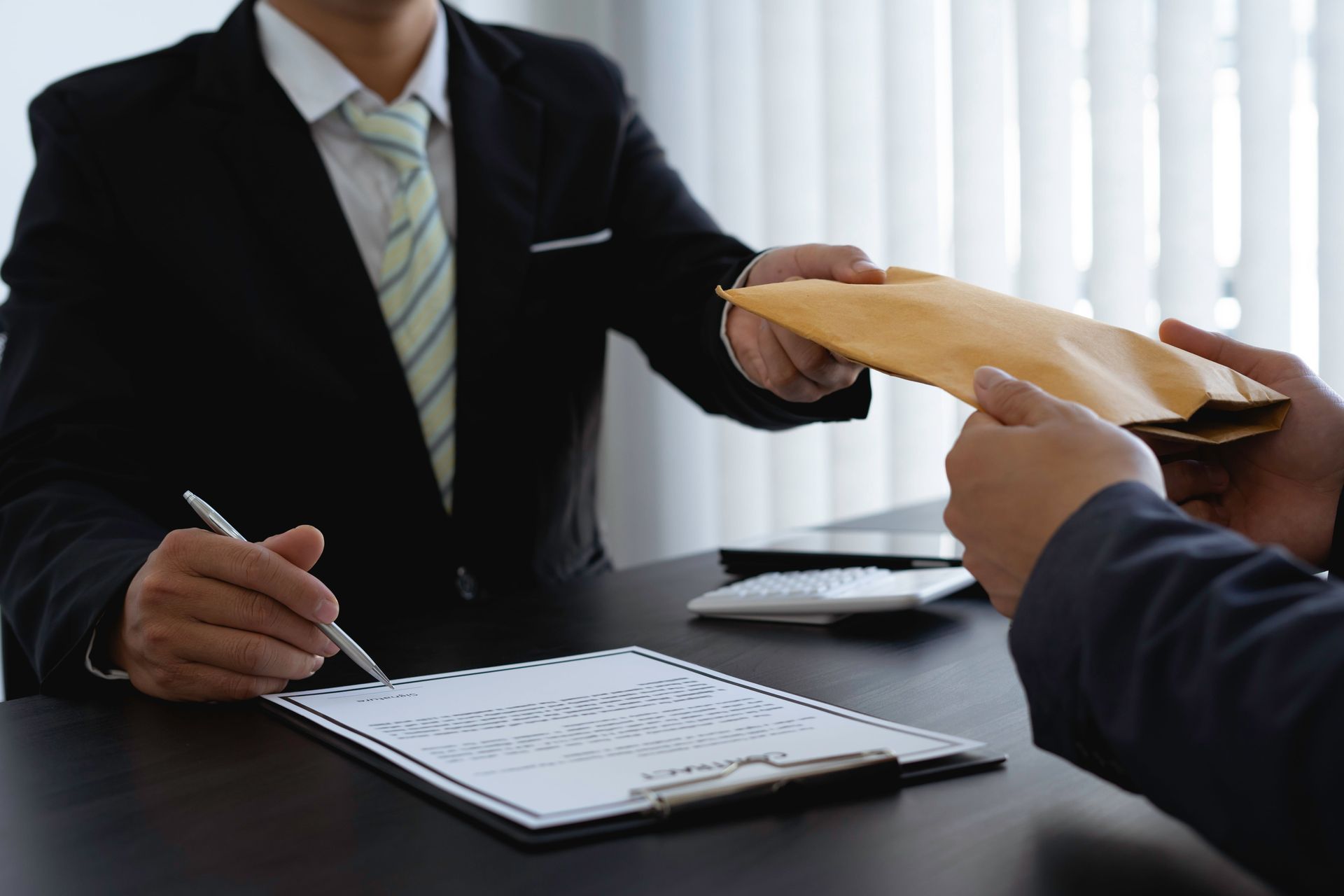 Two men in suits at a desk: one hands an envelope, the other signs a document.