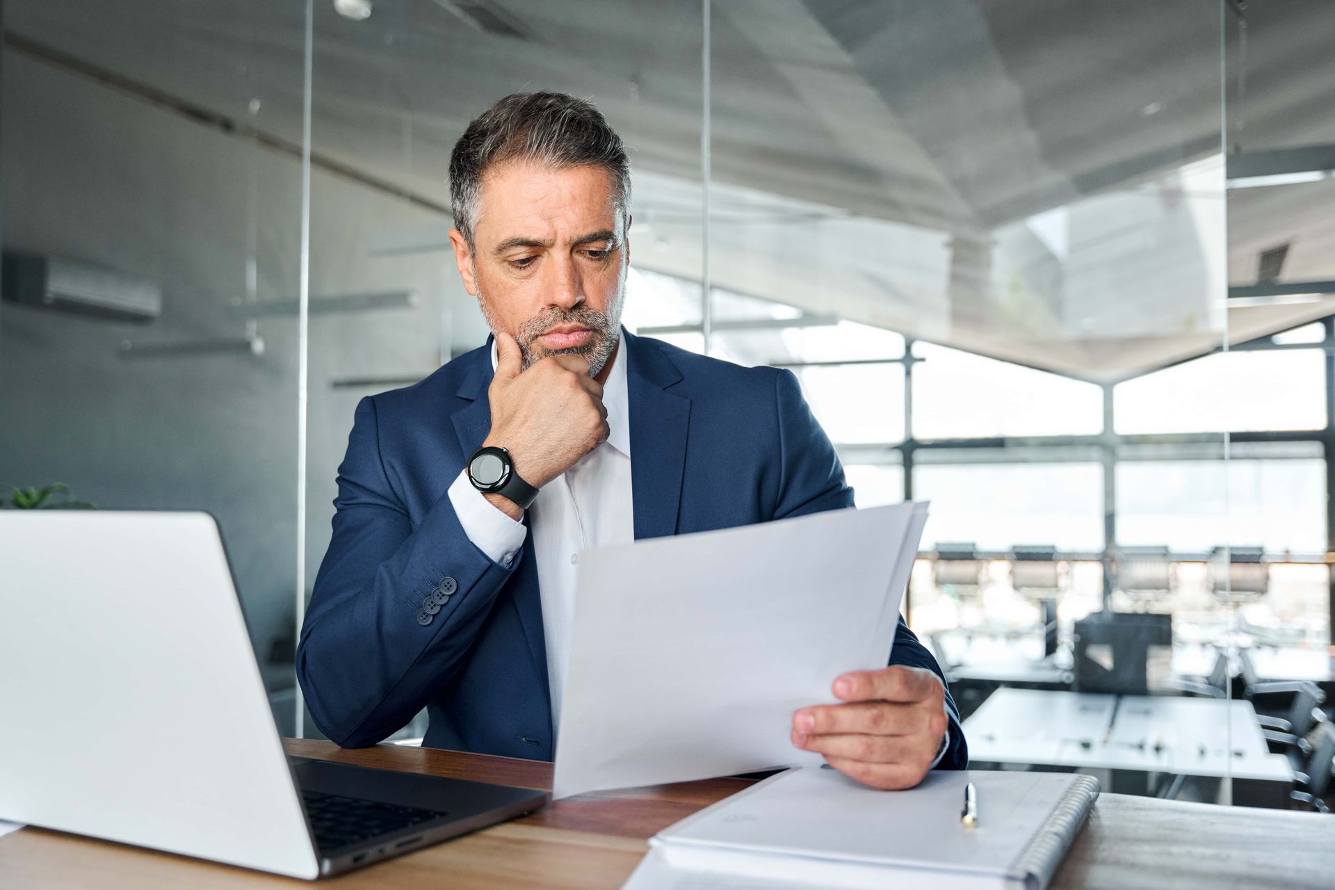 Man in suit reviewing papers, laptop on desk, office setting.