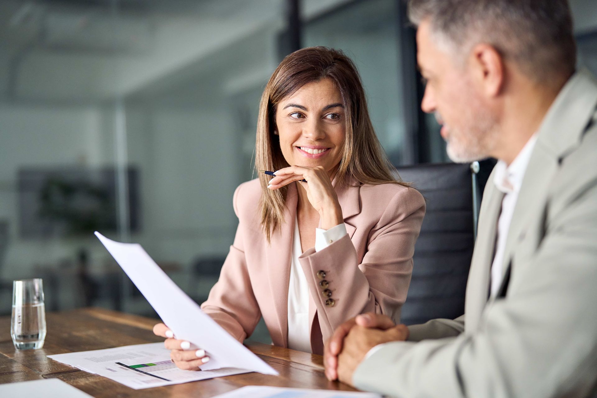 Woman in pink blazer smiles, reviews documents with a man in an office.