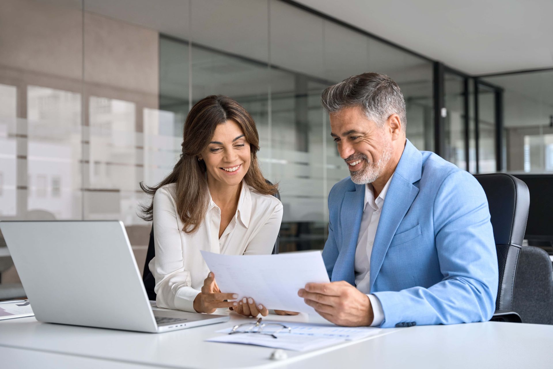 Woman and man reviewing documents, smiling, in an office setting with a laptop.