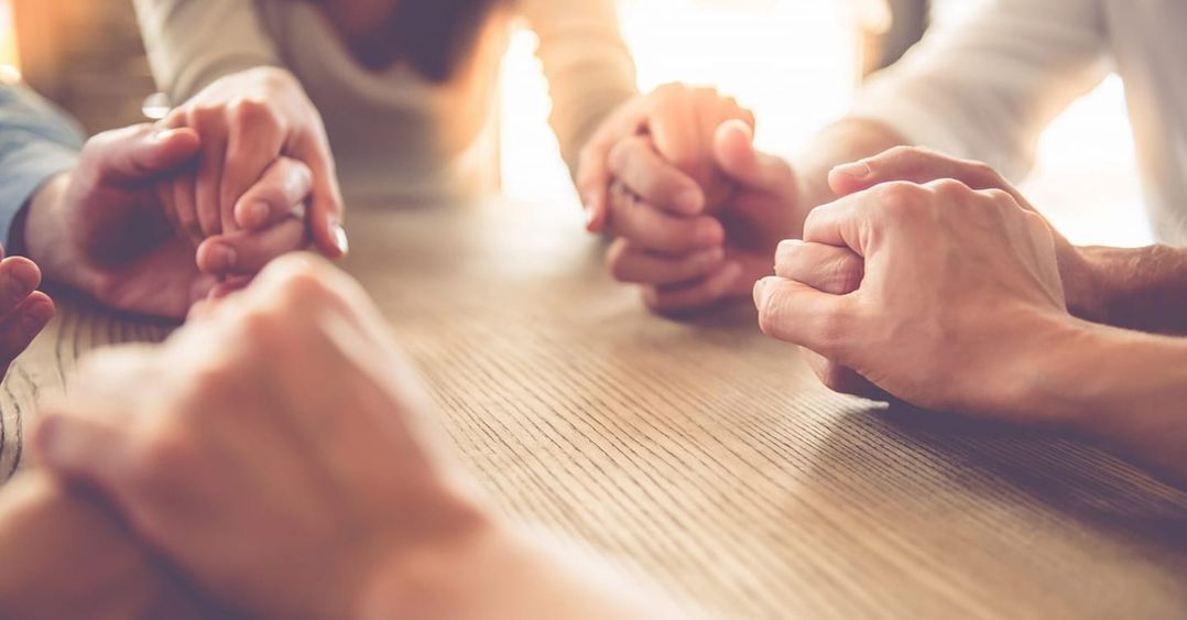 Group of People Are Sitting at a Table With Their Hands Folded in Prayer — Mullumbimby Comprehensive Health Centre in Mullumbimby, NSW