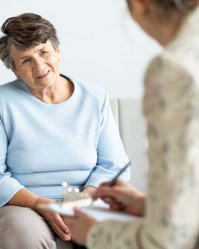Elderly Woman is Sitting on a Couch Talking to a Woman Holding a Clipboard — Mullumbimby Comprehensive Health Centre in Mullumbimby, NSW