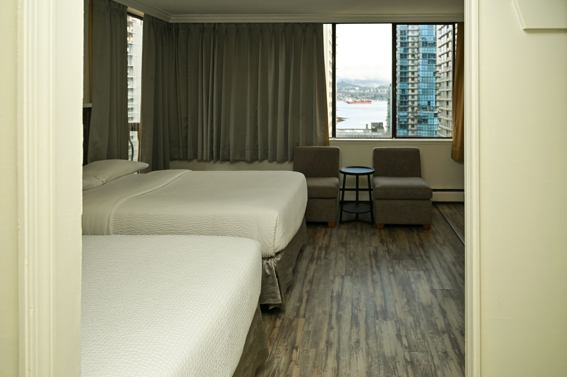 A hotel room with two white beds, two grey chairs, a small table, and a large window looking out onto a city waterfront.