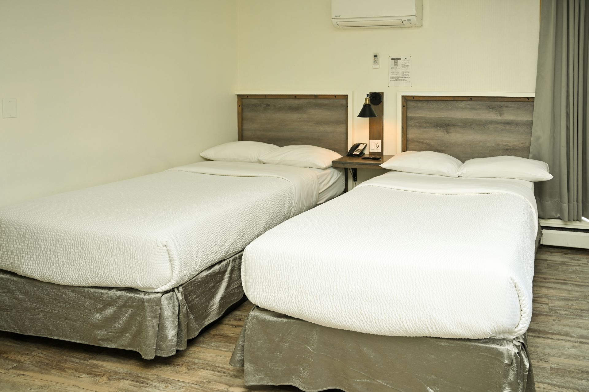 A hotel room with two twin beds featuring white bedding, gray headboards, and wood-patterned flooring.